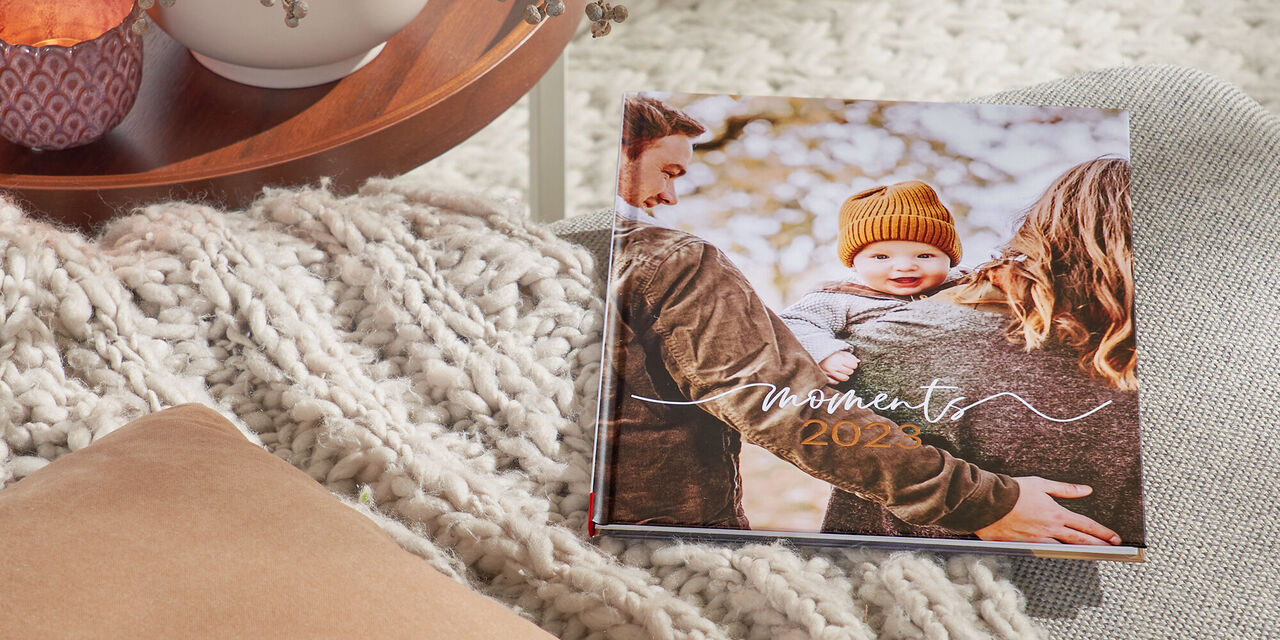 Egy szürke kanapén egy CEWE FOTÓKÖNYV hever. A borítón egy család fotója látható, illetve a "Moments 2023" cím olvasható. Balra van egy asztal teamécsesekkel és egy növénnyel.