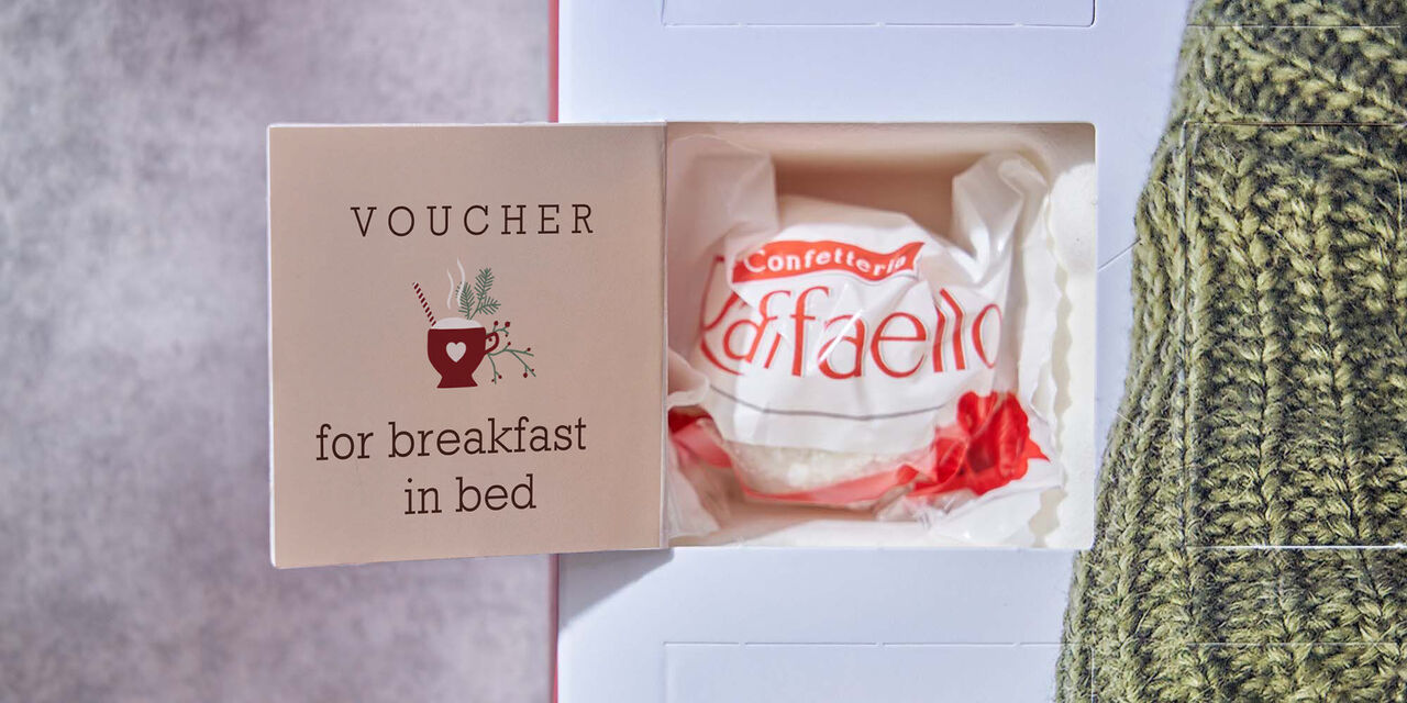 Auf der Innenseite eines geöffneten Adventskalendertürchens steht der Text “Gutschein für Frühstück im Bett”. Außerdem befindet sich eine Tasse mit weihnachtlicher Deko als Designelement auf der Tür, hinter der eine Raffaello-Praline steckt.