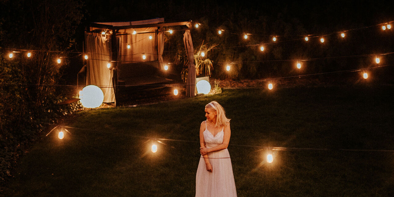 Éjjel a szabadban egy fehér ruhás nő áll. Hangulatos fények veszik körül, a háttérben egy kivilágított pavilon áll.