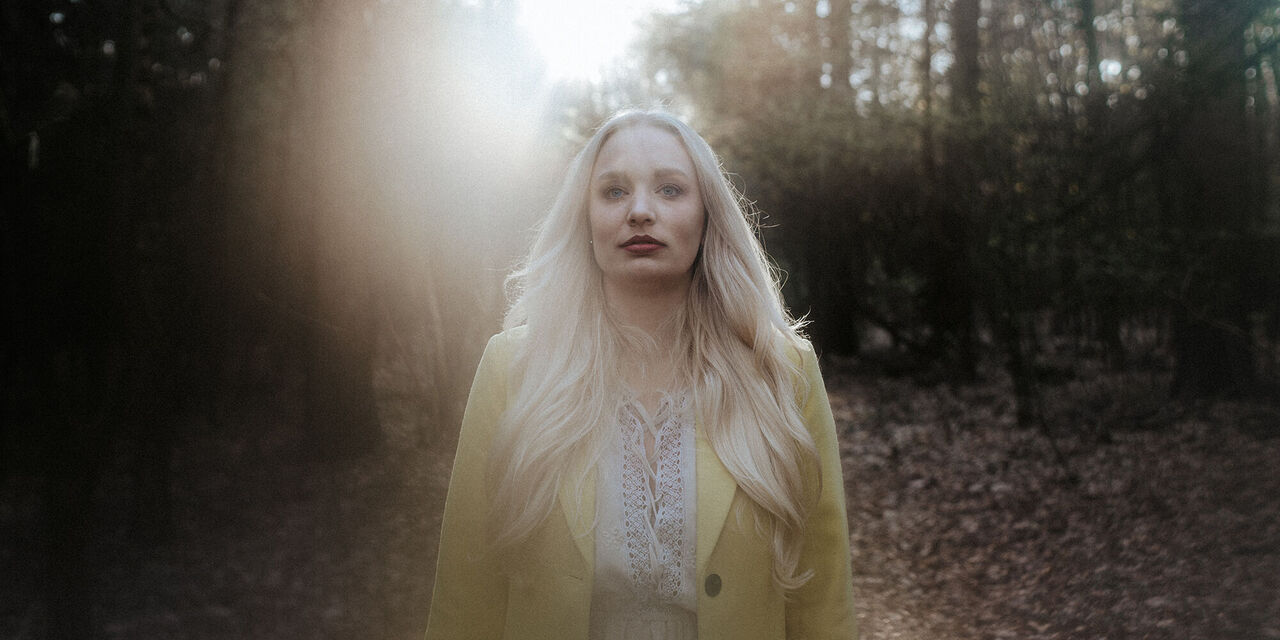 Az erdőben álló, sárga kabátos szőke hölgyre hátulról vetődnek a napsugarak, a háttér enyhén elmosódott.
